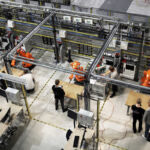 en fabriksgolv med maskiner och arbetare i orangea västar